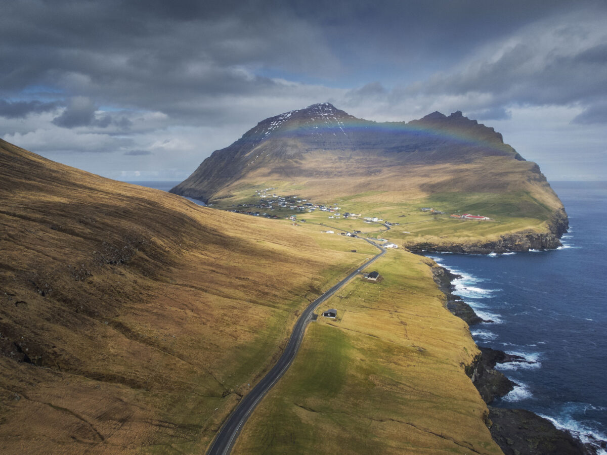 Aerial view of Viðareiði village on Viðoy Island, Faroe Islands, with winding coastal road and rainbow over dramatic mountains and ocean.