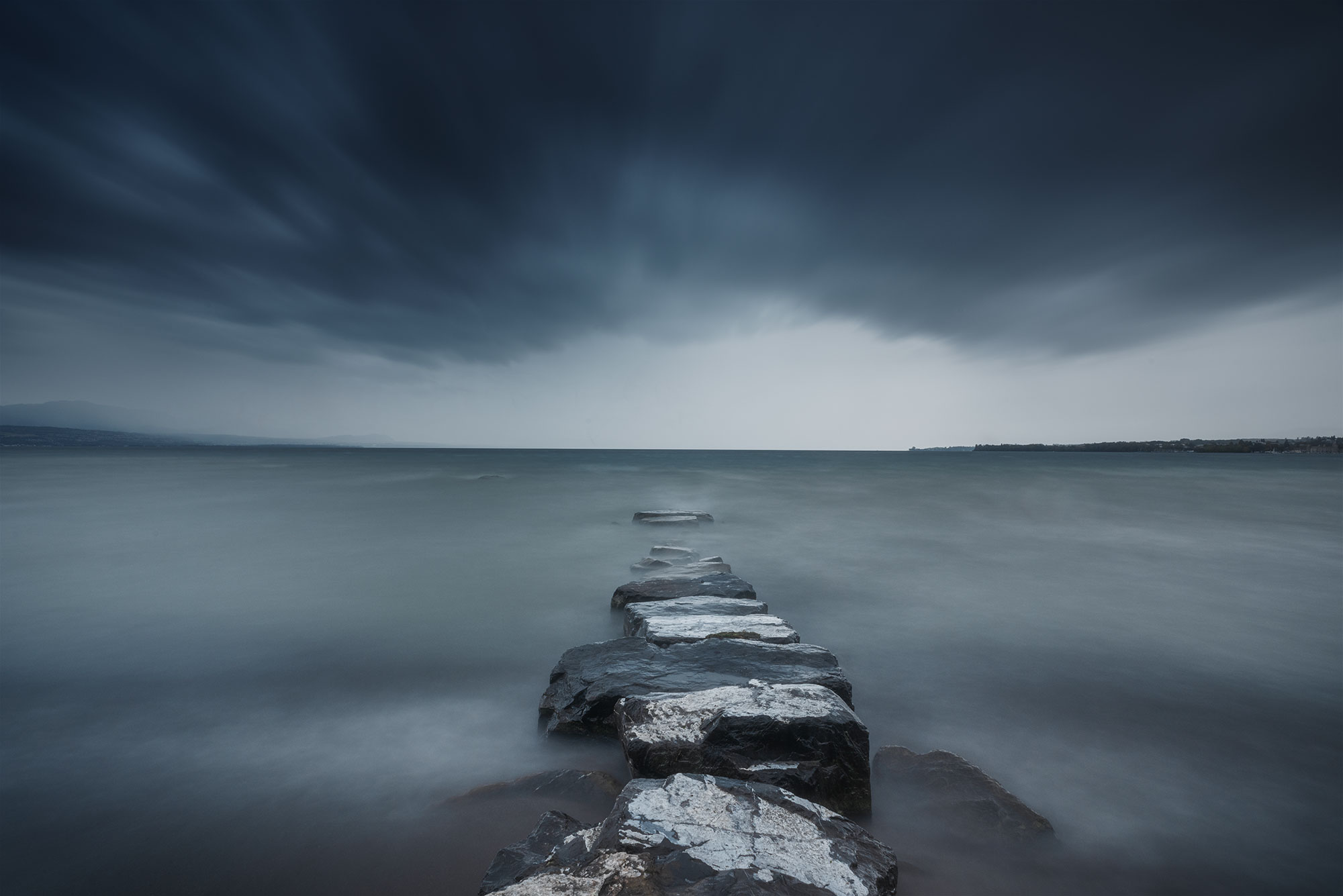This compelling image encapsulates the dynamic beauty of Préverenges Beach in Switzerland during a storm, as captured through a long exposure landscape photograph. The turbulent weather adds an element of drama to the scene, with the dark, moody clouds swirling above Lake Geneva. The extended exposure smoothens the water's surface, creating a surreal reflection of the stormy atmosphere. The contrast between the agitated sky and the serene lake serves as a poignant reminder of nature's power and beauty. This photograph invites viewers to appreciate the intensity of the moment and the unique perspective offered by long exposure photography.