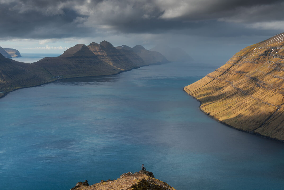 Hiker overlooking fjord from Klakkur viewpoint on Borðoy Island, Faroe Islands, with dramatic mountains and soft light.