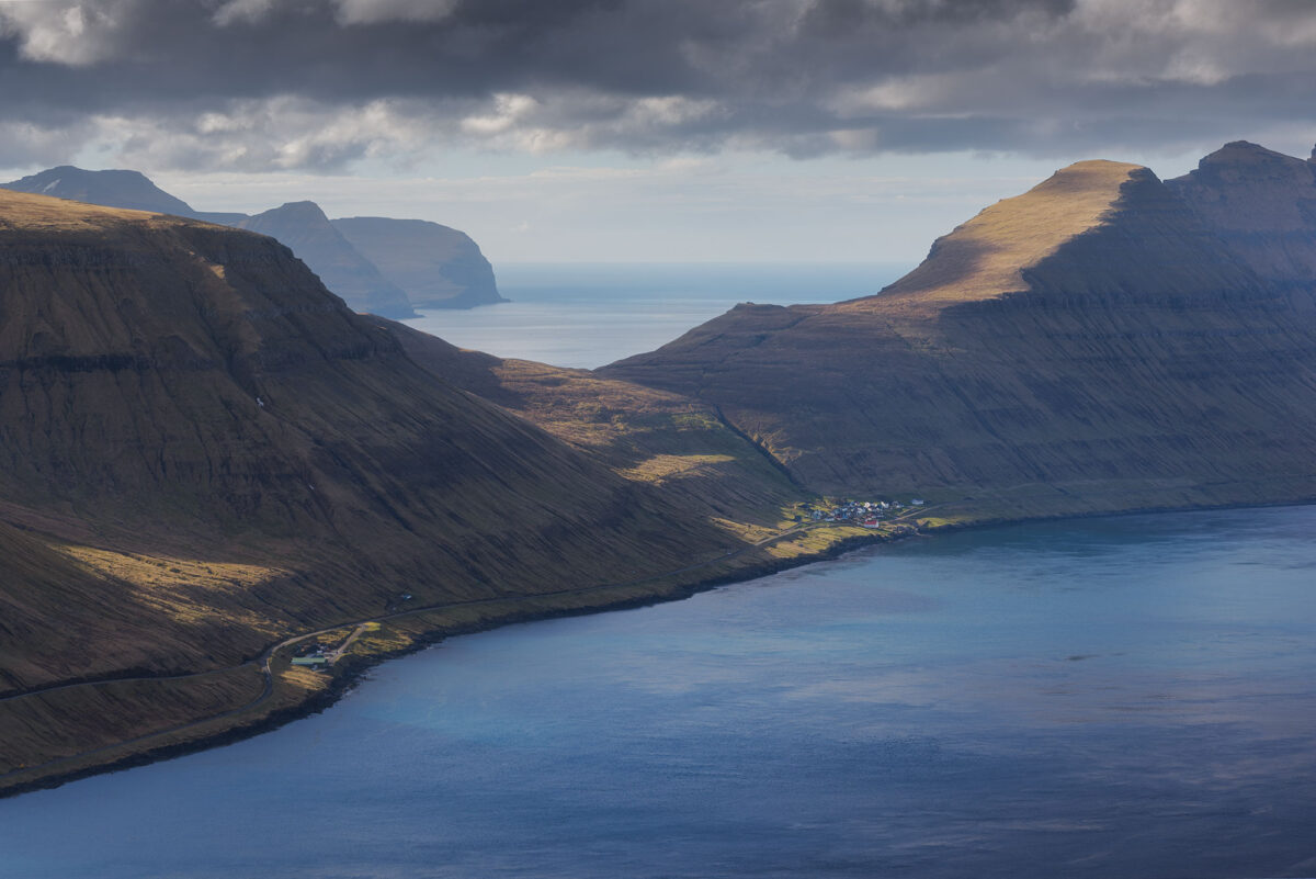 View over fjord and coastal village from Klakkur viewpoint, Faroe Islands, with layered mountains and moody sky.