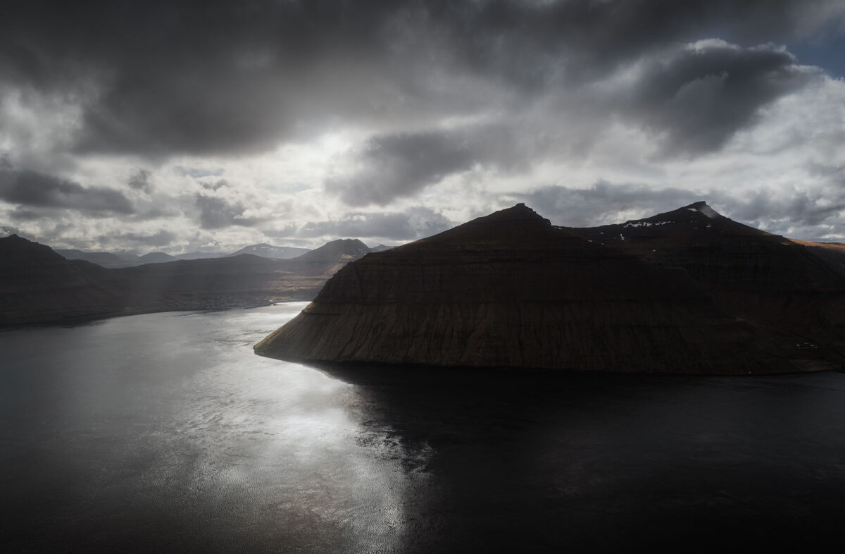 Sunlight breaking through clouds over fjord at Klakkur, Faroe Islands, creating reflections on the water and dramatic shadows.