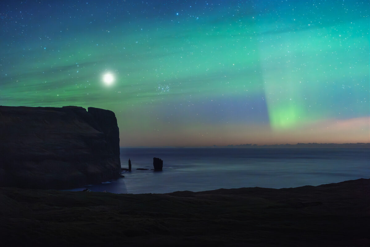Northern lights and airglow above Risin og Kellingin sea stacks, Faroe Islands, under a bright full moon and starry sky.