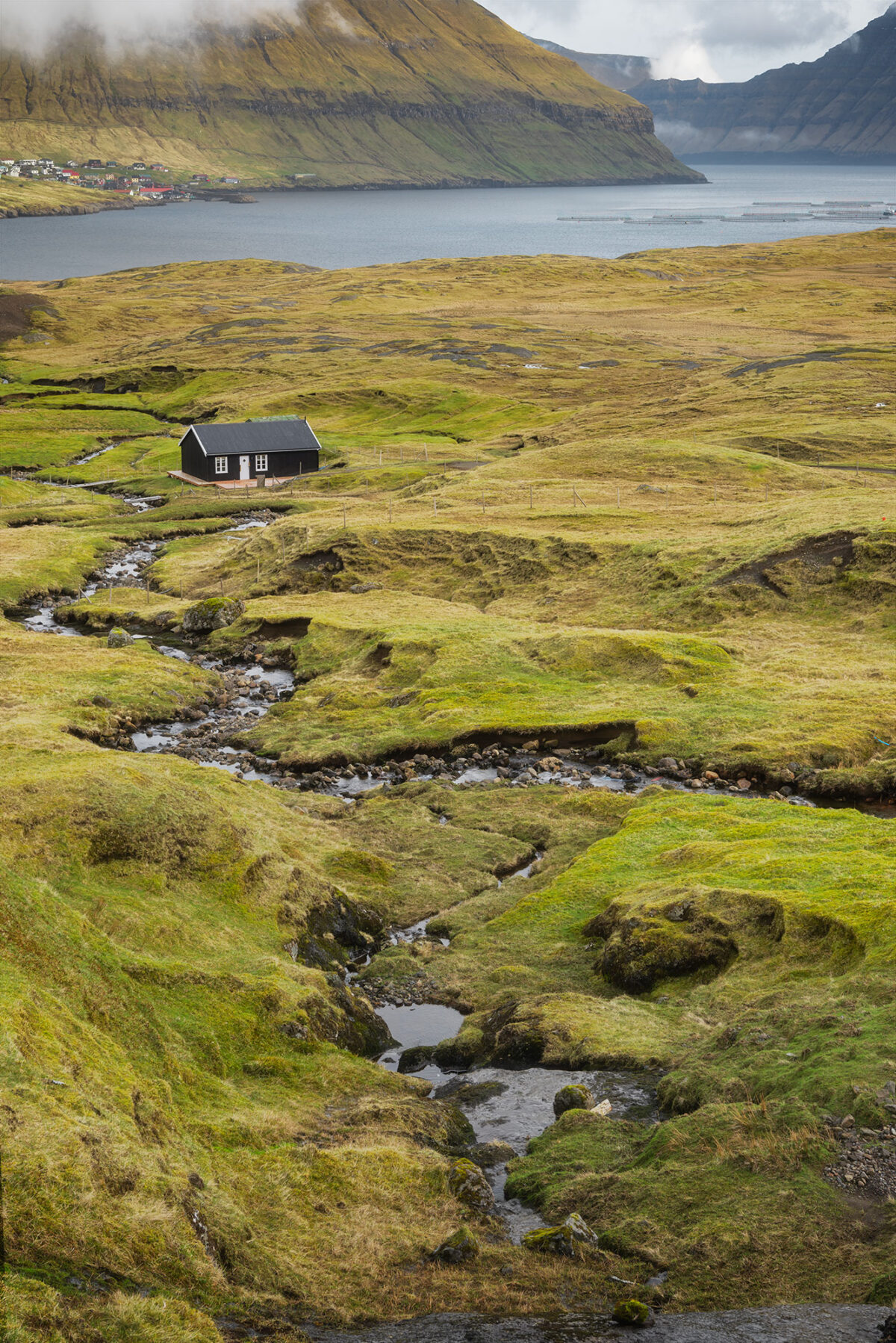 Black house in Hellur, Faroe Islands, with winding stream leading through green landscape toward fjord and mountains