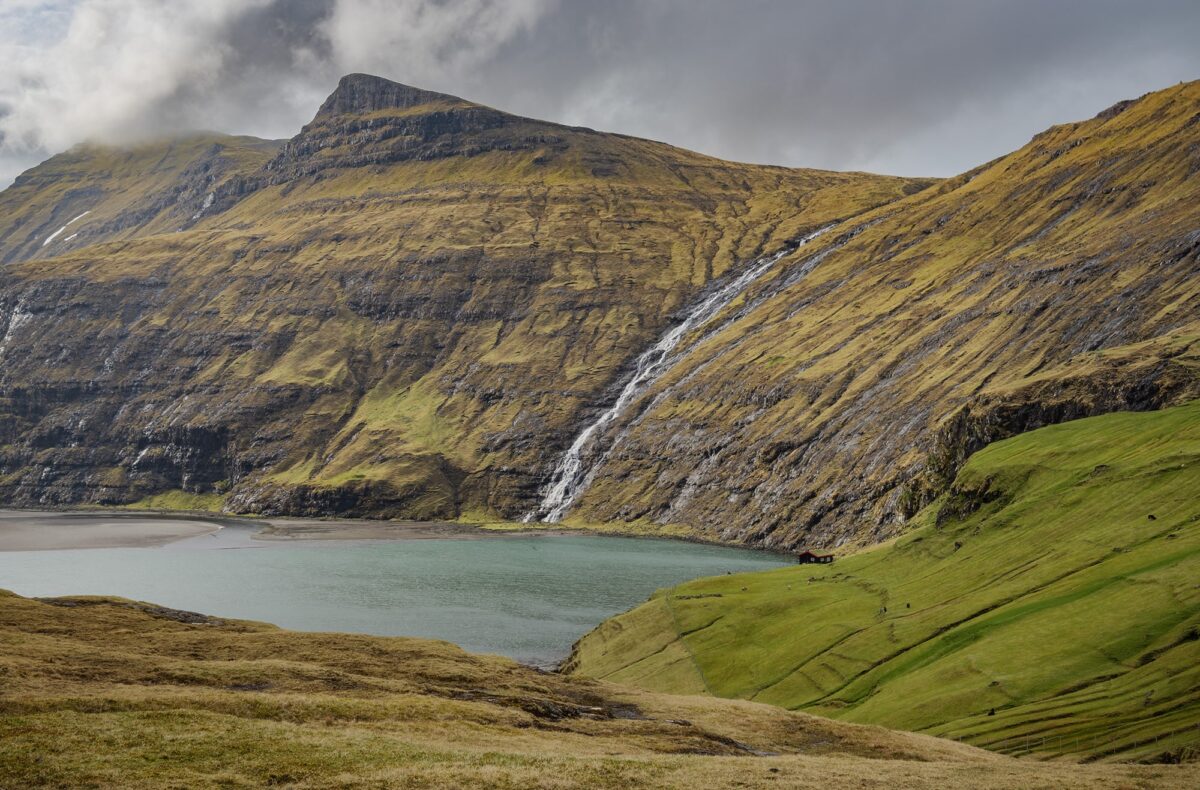 View of Saksun lagoon with waterfall cascading down the mountains and a small red house on the hillside, Faroe Islands.
