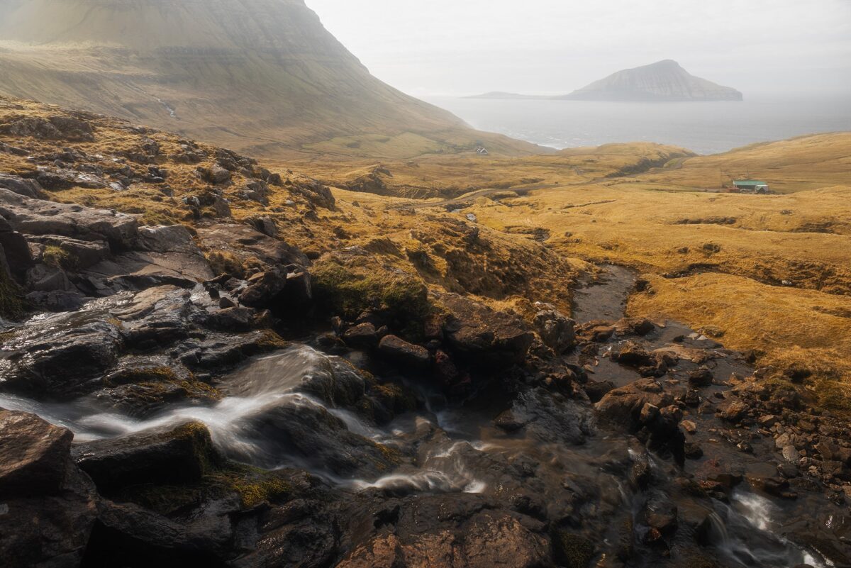Small waterfall flowing through rocky landscape above Norðradalur, Faroe Islands, overlooking fjord and Koltur island.