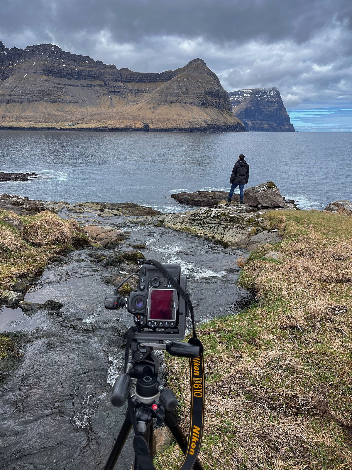 This exhilarating image captures the thrill of the moment as my Nikon D810 frames a stunning scene on Viðoy Island. With the powerful lens focused on the cascading waterfall, the camera immortalizes the daring adventure of my friend bravely standing below the rushing waters.