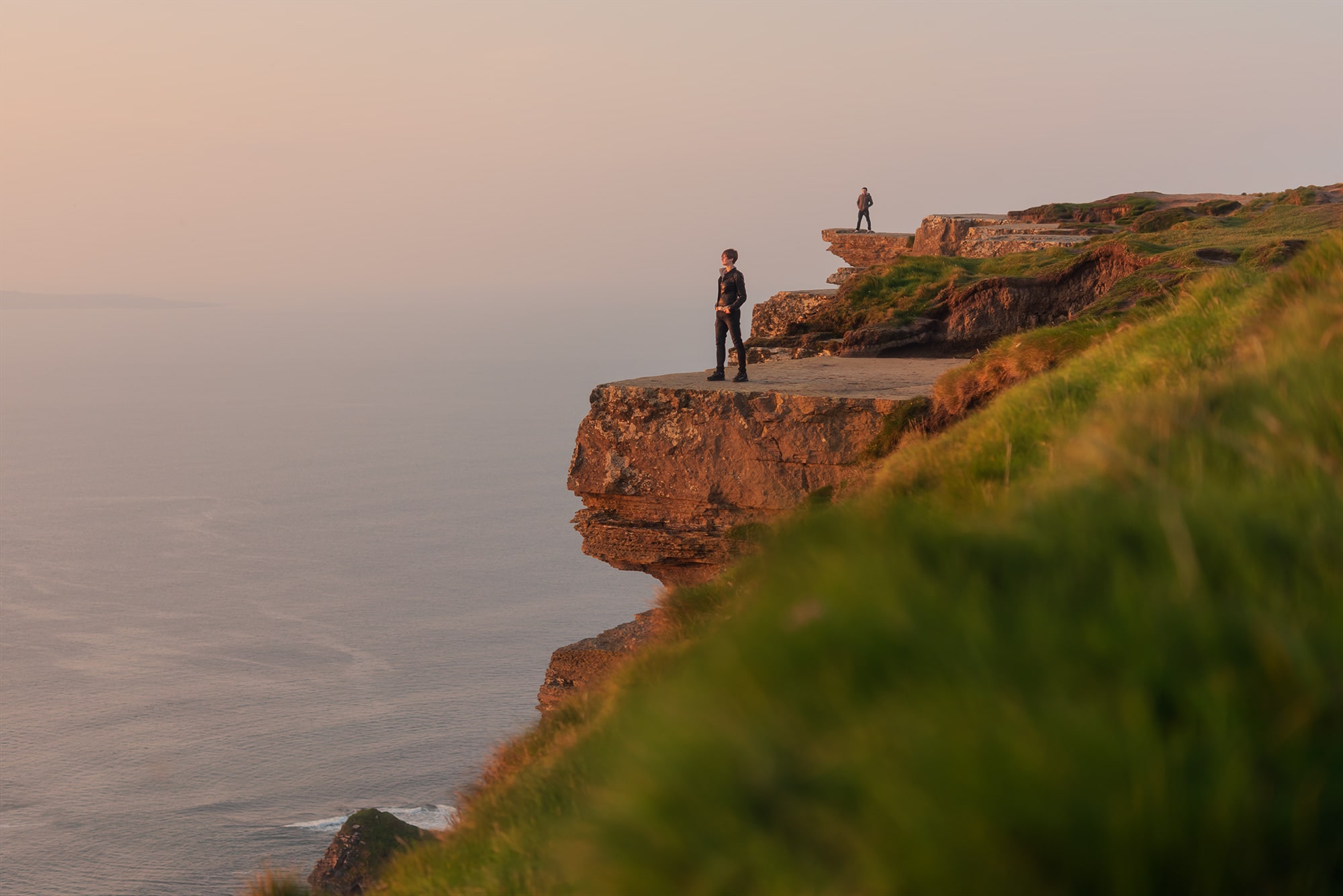 This stunning landscape photography showcases the iconic cliffs at sunset, bathed in the warm hues of the evening sky. Amidst this awe-inspiring scenery, two figures stand on the cliffs, providing a sense of scale and perspective to the vastness of the landscape. Join Jennifer Esseiva on this visual journey as she captures the magic of one of Ireland's most renowned landmarks, inviting viewers to immerse themselves in the timeless beauty of the Cliffs of Moher.