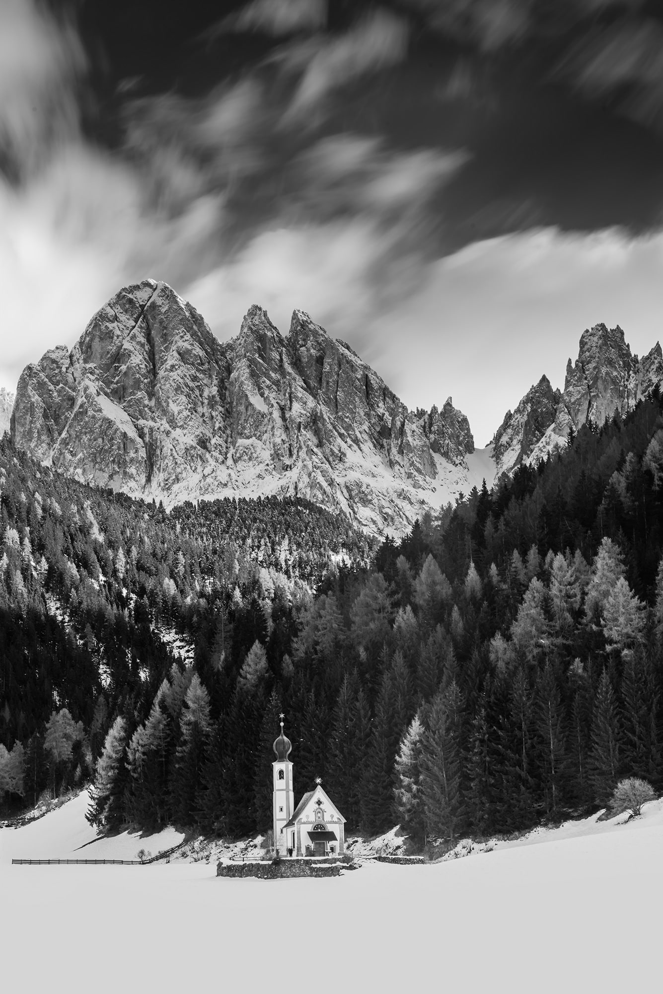Experience the striking contrast of light and shadow in this dramatic black and white landscape photograph of the Chiesetta di San Giovanni in Ranui, nestled in the heart of the Dolomites. Captured by renowned photographer Jennifer Esseiva with her Nikon D810, this image showcases the timeless beauty of the iconic church against the rugged backdrop of the mountains. The monochromatic tones add depth and intensity to the scene, highlighting the intricate details of the architecture and the surrounding landscape. Jennifer Esseiva's skillful composition and mastery of black and white photography invite viewers to immerse themselves in the timeless allure of this iconic location in the Dolomites.