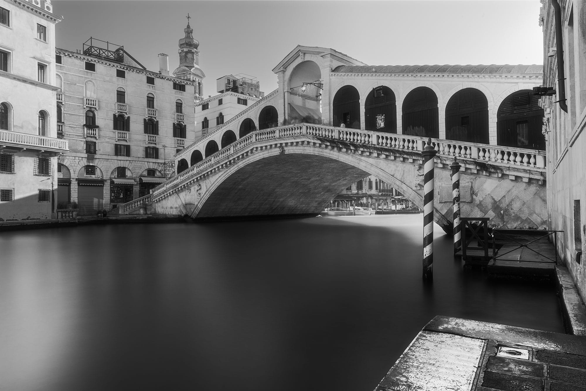 Experience the timeless allure of Venice's Rialto Bridge in this stunning black and white long exposure photograph. Captured with minimalist precision by travel photographer Jennifer Esseiva using her Nikon D810, this image exudes a sense of elegance and tranquility. The smooth waters of the canal create a striking contrast against the architectural marvel of the bridge, making it an ideal addition to elevate any office space. Let the iconic beauty of Venice inspire productivity and creativity with this captivating landscape photography masterpiece.
