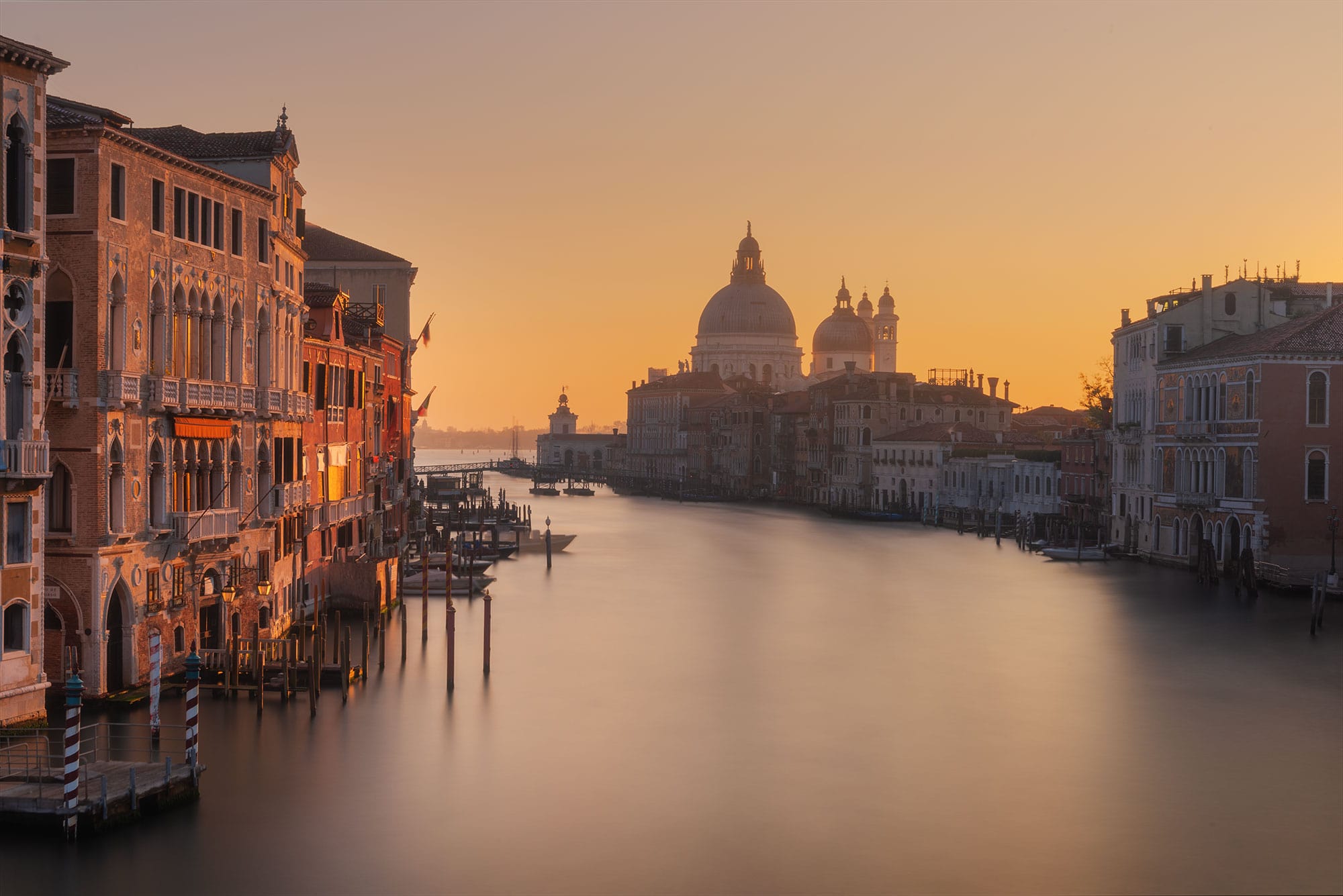 Experience the enchanting beauty of Venice at sunrise with this captivating urban landscape photograph. The iconic Grand Canal and Basilica Santa Maria della Salute are bathed in warm hues of orange, creating a mesmerizing scene that evokes a sense of tranquility and wonder. Taken by the skilled eye of a female travel photographer in Italy, this stunning image captures the essence of Venice's timeless charm and architectural splendor. Immerse yourself in the romantic allure of the Grand Canal as it comes to life in the soft light of dawn, beautifully captured by the talented photographer.