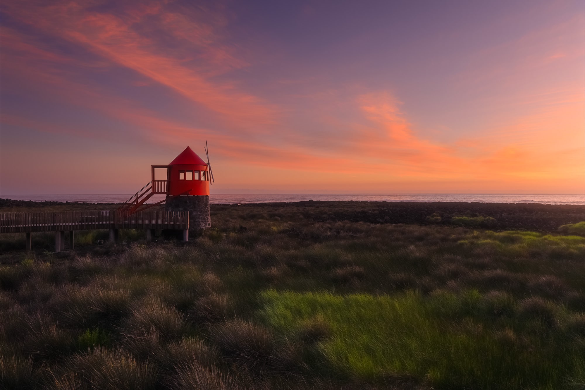 Experience the breathtaking beauty of a sunset at Lajes do Pico in the Azores through the lens of landscape photographer Jennifer Esseiva and her Nikon D810. In this captivating image, the iconic red windmill stands as a silhouette against the vibrant hues of the pink sky, casting a serene glow over the tranquil seascape below. With her expert composition and keen eye for detail, Esseiva captures the essence of this picturesque moment, inviting viewers to immerse themselves in the enchanting beauty of the Azores. Join Esseiva on this visual journey, where every frame is a testament to the timeless allure of nature's grandeur. Perfect for travel bloggers and enthusiasts seeking to capture the magic of the Azorean sunset.