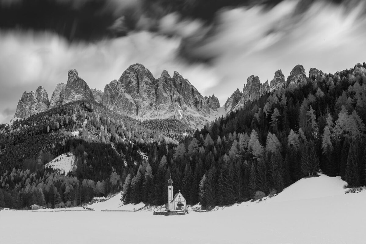 Immerse yourself in the captivating interplay of light and shadow with this striking black and white landscape photograph of the Chiesetta di San Giovanni in Ranui, nestled amidst the Dolomites. Renowned photographer Jennifer Esseiva expertly captured this scene with her Nikon D810, showcasing the enduring charm of the iconic church set against the rugged mountain backdrop. The monochromatic tones heighten the sense of depth and drama, accentuating the intricate architectural details and the surrounding landscape. Esseiva's masterful composition invites viewers to journey into the timeless beauty of this iconic Dolomite location.