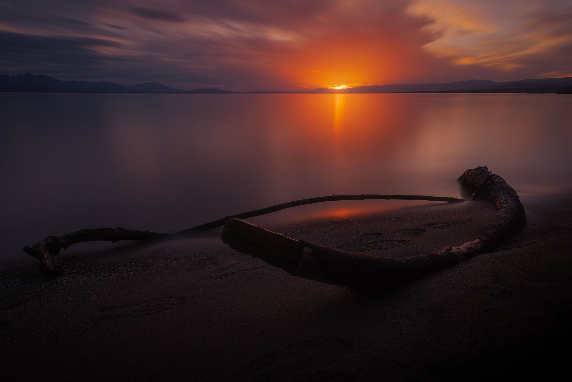 Embark on a visual journey through the lens of landscape photographer Jennifer Esseiva as she skillfully captures the breathtaking beauty of a long exposure dramatic sunset over Lake Geneva in St. Sulpice, Canton of Vaud, Switzerland. The lake shore, adorned with weathered wood pieces on the sand, adds a rustic charm to the scene. Jennifer's photography transforms the sky into a canvas of vibrant hues, creating a stunning visual spectacle. Immerse yourself in the dynamic allure of this Swiss sunset, expertly captured to evoke a sense of awe and wonder.