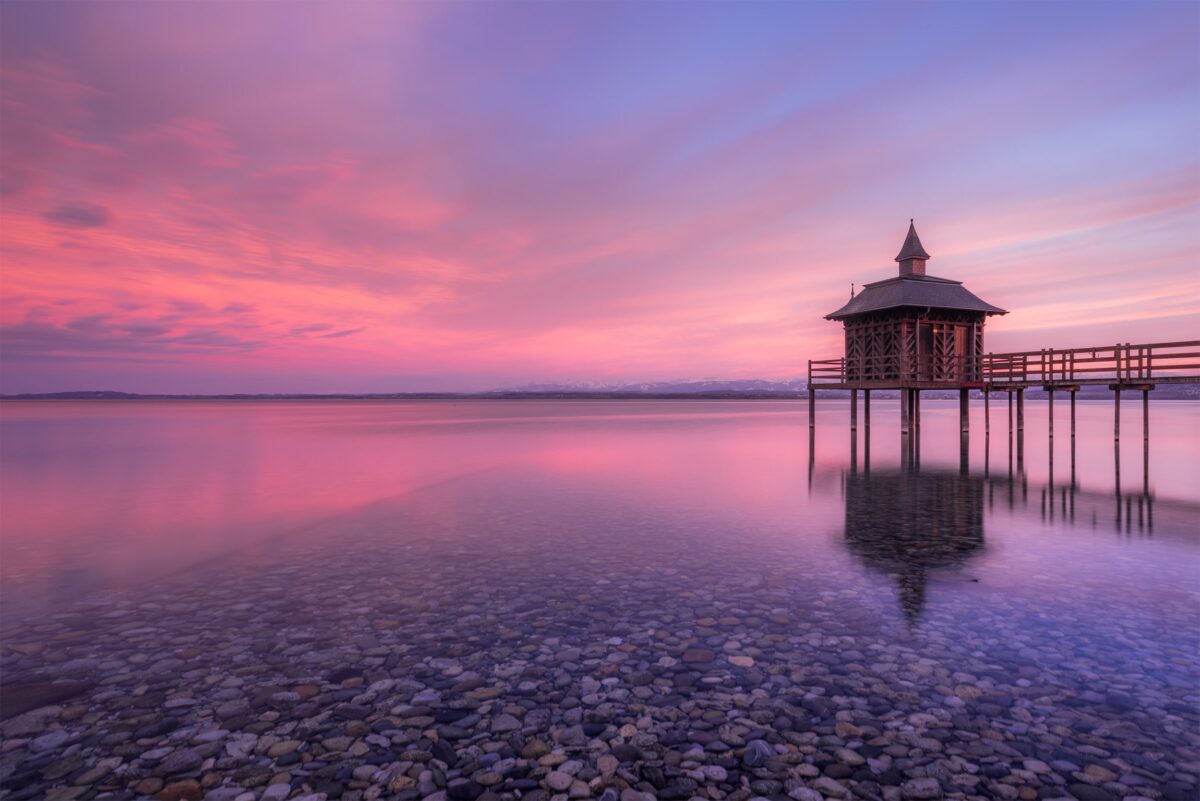 Long exposure photography of Lake Neuchâtel in Switzerland at sunset in winter, featuring a wooden pavilion on calm water with soft pink sky reflections