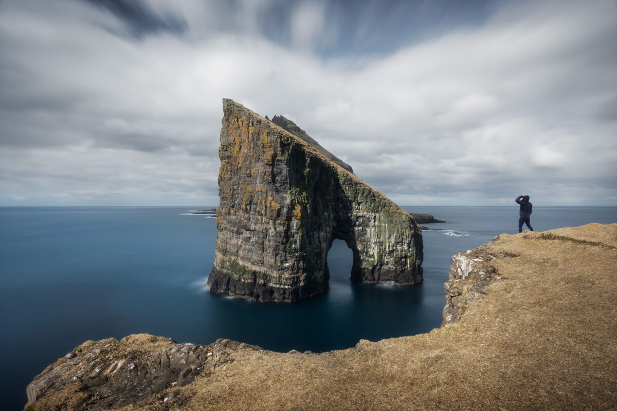 Person overlooking Drangarnir sea stacks from Vágar Island, Faroe Islands, in long exposure coastal landscape.