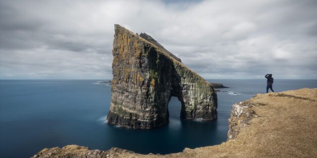 Experience the captivating allure of the Faroe Islands with stunning landscape photography, featuring a mesmerizing long exposure shot capturing a lone figure gazing upon the majestic Drangarnir Sea Stacks from Vágar Island. Immerse yourself in the dramatic beauty of this remote seascape, rendered in exquisite detail through the lens of our photographer.