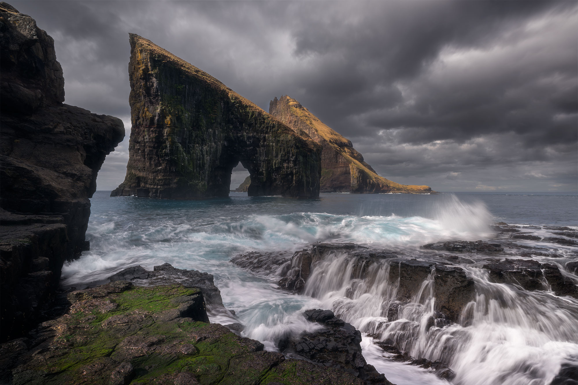Indulge in the mesmerizing beauty of the Faroe Islands with captivating landscape photography, showcasing the iconic Drangarnir sea stacks as seen from Vágar Island. This stunning long exposure shot, expertly captured by photographer Jennifer Esseiva, brings to life the rugged charm and dramatic allure of this remote seascape.