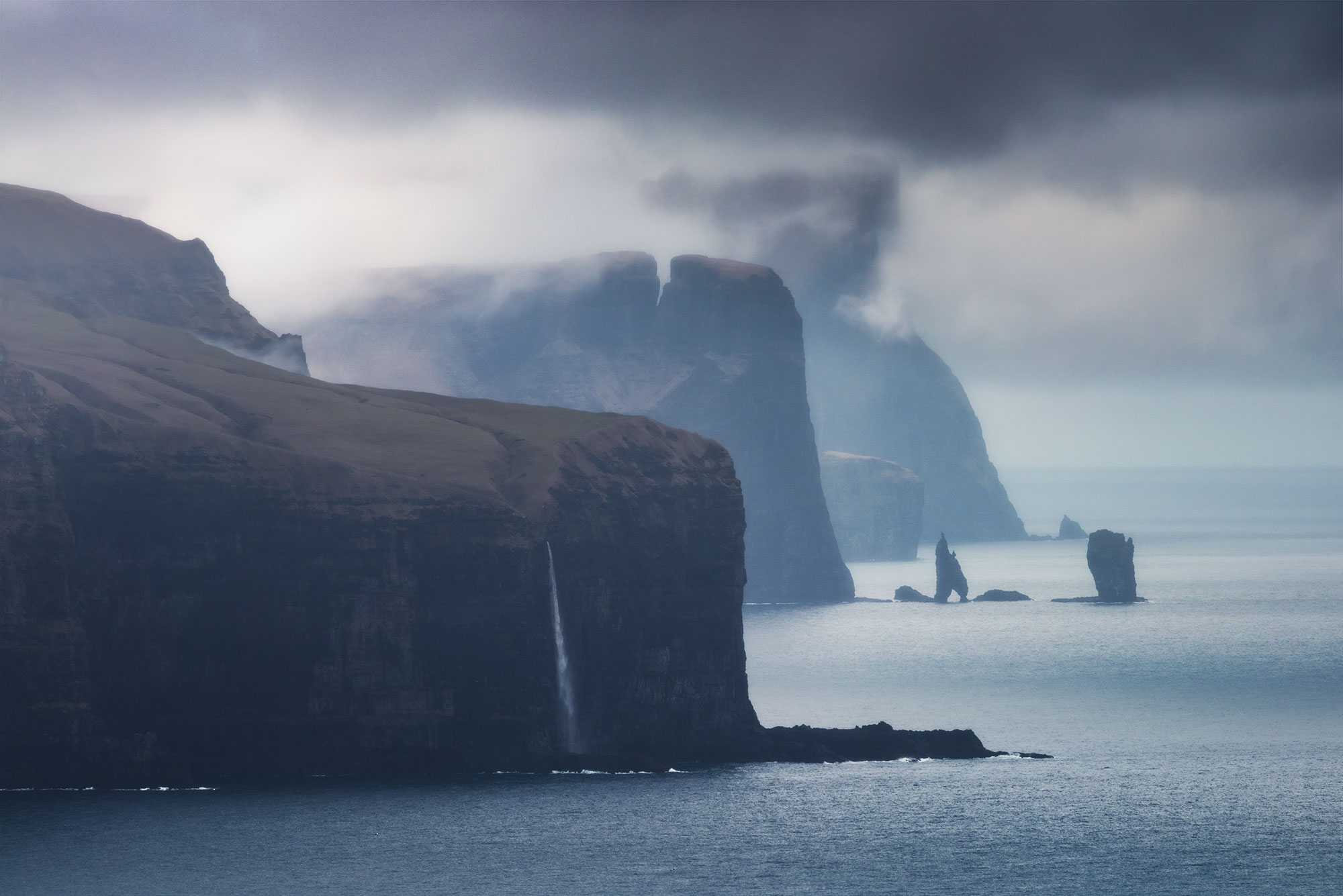 Step into the mystical world of the Faroe Islands with this captivating landscape photograph captured from Kalsoy. Behold the misty and moody vista, where a waterfall cascades into the ocean amidst the dramatic backdrop of the two iconic sea stacks, Risin & Kellingin. This mesmerizing image encapsulates the raw beauty and rugged charm of the Faroe Islands, inviting viewers to immerse themselves in its ethereal allure. Experience the magic of nature's drama unfolding through the lens of this evocative landscape photograph. Explore the timeless allure of the Faroe Islands in this captivating portrayal of its mystical landscapes.