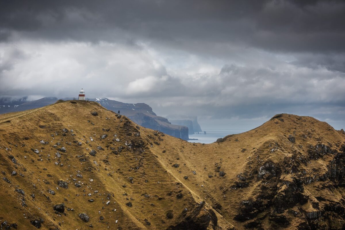 Kallur Lighthouse on Kalsoy Island, Faroe Islands, with person on cliffs under dramatic cloudy sky overlooking ocean.