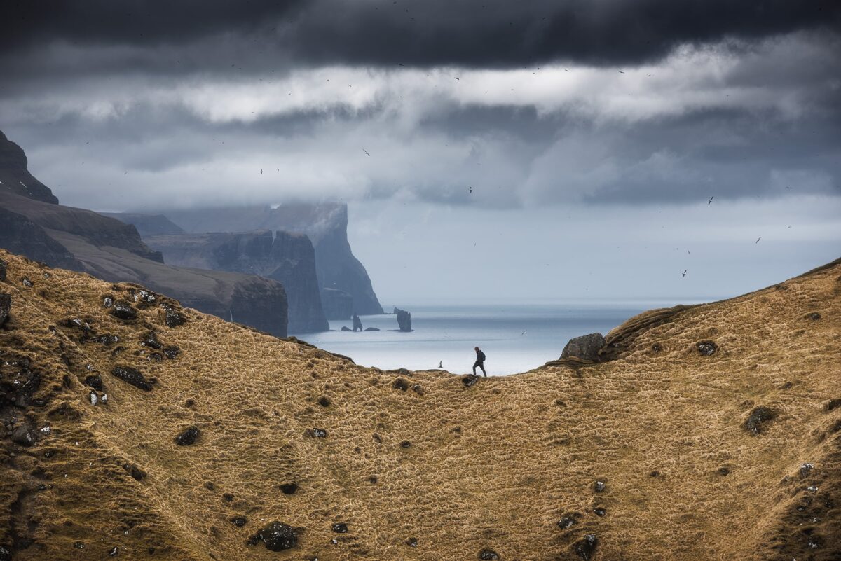 Kallur Lighthouse on Kalsoy Island, Faroe Islands, with person on cliffs under dramatic cloudy sky and ocean views.