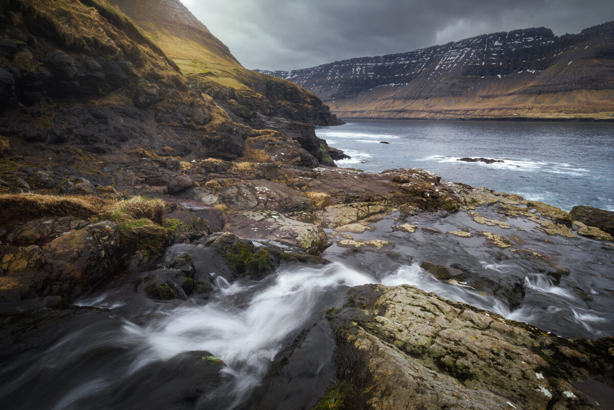 Long exposure of river flowing from Viðareiði village to the ocean on Viðoy Island, Faroe Islands, with rugged coastal landscape.