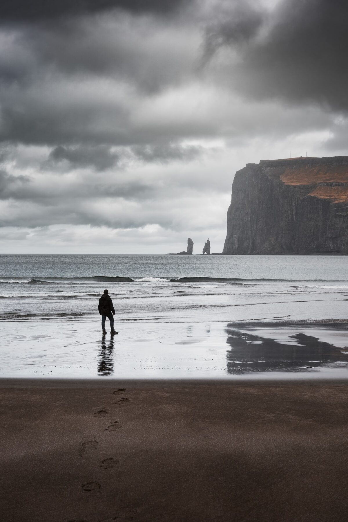 Person on black sand beach of Tjørnuvík, Faroe Islands, looking toward Risin and Kellingin sea stacks with waves and cliffs.