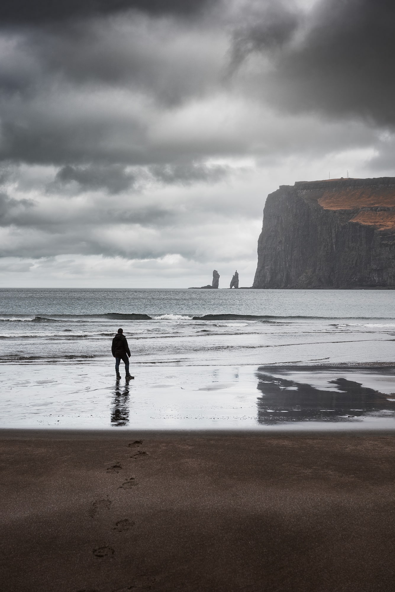 Embark on a visual journey through breathtaking landscape photography, featuring a captivating image of a man gazing upon the two sea stacks from the black sand beach of Tjørnuvík, nestled in the northernmost village of Streymoy island in the Faroe Islands. Immerse yourself in the rugged beauty of this remote location as the waves crash against the shoreline, creating a mesmerizing scene against the dramatic cliffs. Witness the harmonious blend of nature's elements captured in this evocative composition.