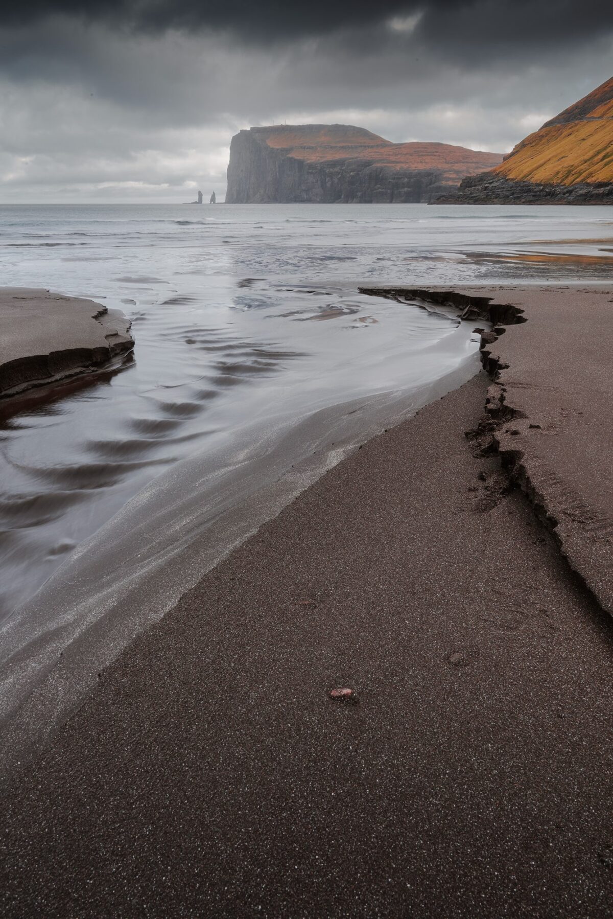 Risin and Kellingin sea stacks seen from black sand beach of Tjørnuvík, Faroe Islands, with rugged cliffs and ocean waves.