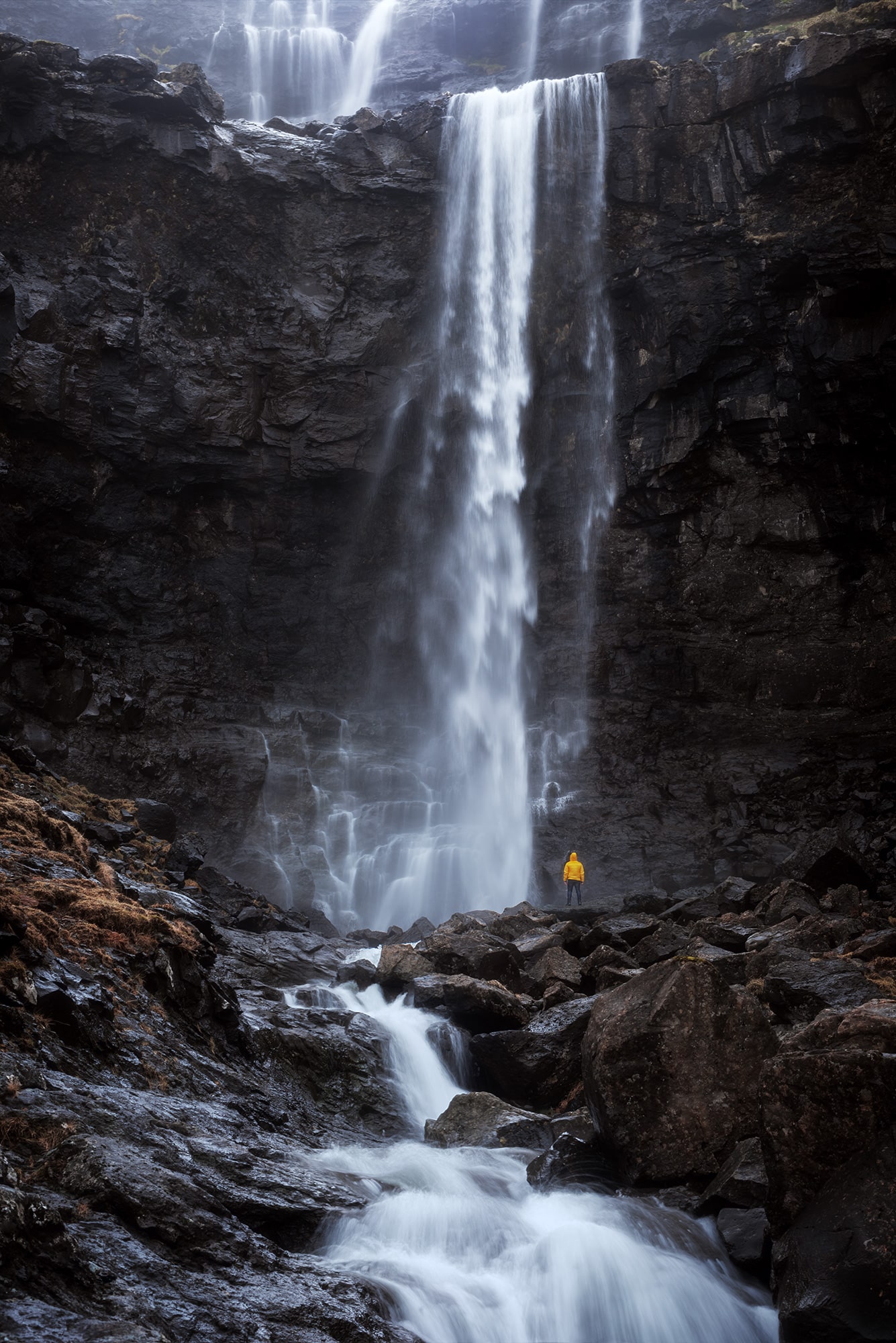 Embark on a visual journey through mesmerizing landscape photography capturing a lone figure clad in a vibrant yellow raincoat standing at the base of the majestic Fossa waterfall in the Faroe Islands. Marvel at the sheer magnitude and natural beauty as the cascading water plunges into the rugged terrain, creating an awe-inspiring spectacle. Immerse yourself in the raw power of nature and the serene tranquility of this remote island landscape.
