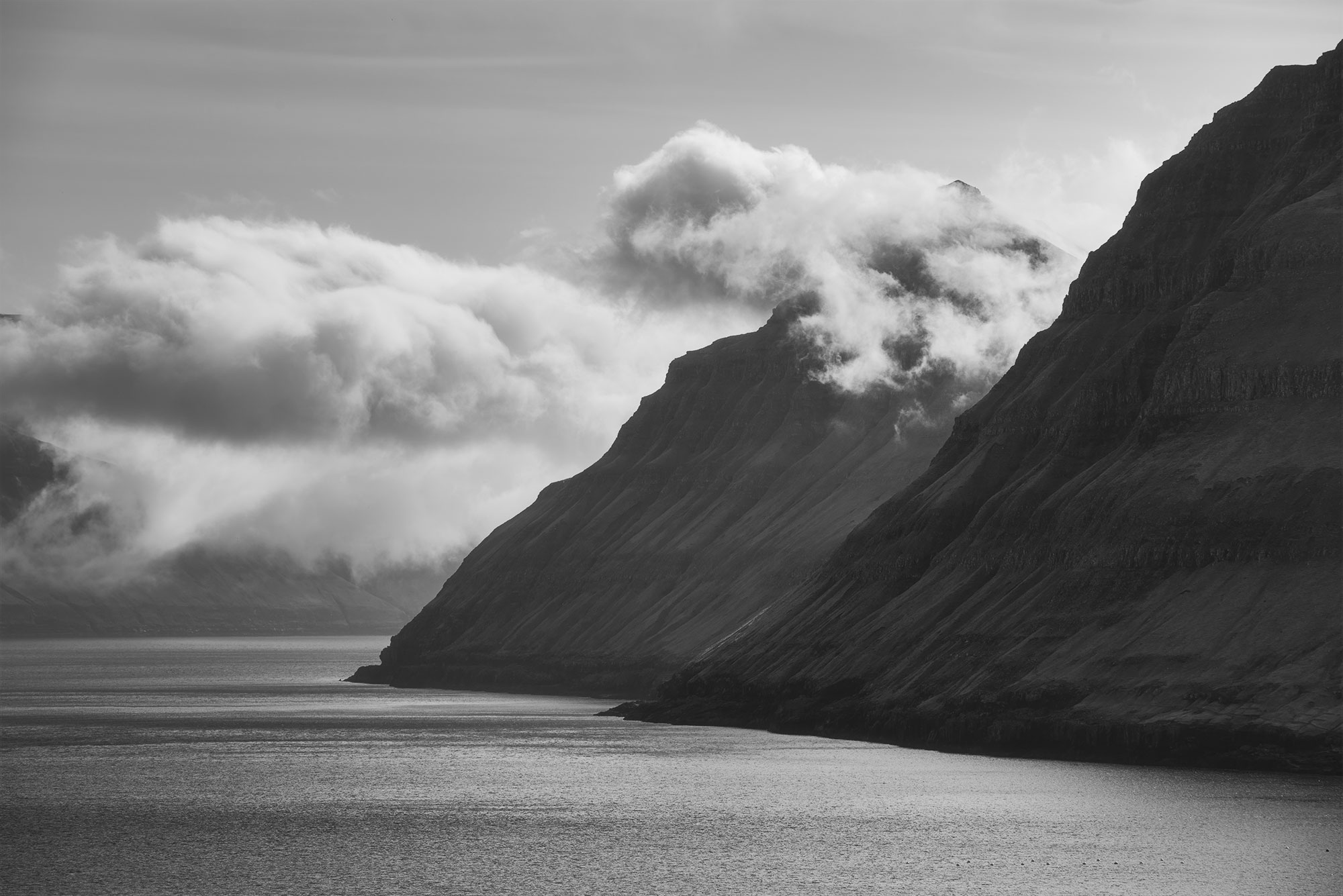 Explore the dramatic landscapes of the Faroe Islands with this captivating black and white photograph captured by Swiss landscape photographer Jennifer Esseiva and her Nikon D810. Behold the rugged beauty of Eysturoy Island as a cloud-covered mountain rises majestically into the sky. This striking image captures the raw, untamed essence of Faroese scenery, inviting viewers to immerse themselves in its timeless allure. Experience the power and majesty of nature preserved in black and white as you delve into this mesmerizing landscape photograph. Discover the unique beauty of the Faroe Islands through Jennifer Esseiva's lens, where every frame tells a story of nature's grandeur.