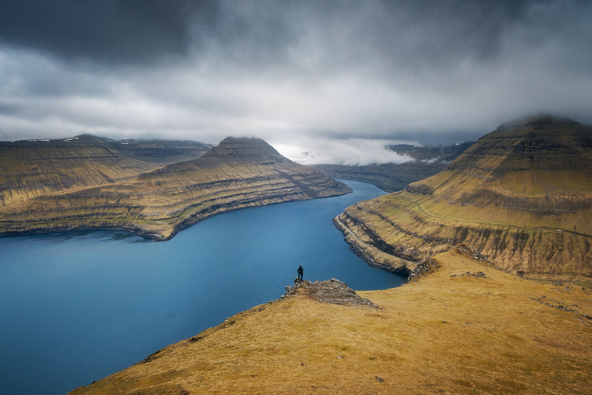 Immerse yourself in the breathtaking landscapes of the Faroe Islands with this captivating landscape photograph captured by Jennifer Esseiva and her Nikon D810. Witness the awe-inspiring scene as a man stands on the edge of a cliff, admiring the dramatic beauty of the Faroe Islands. This striking image captures the rugged charm and untamed wilderness of the archipelago's landscapes, inviting viewers to explore its serene allure. Experience the magic of Faroese scenery through Jennifer Esseiva's lens, where every frame tells a story of nature's grandeur. Discover the timeless beauty of the Faroe Islands in this evocative landscape photograph.