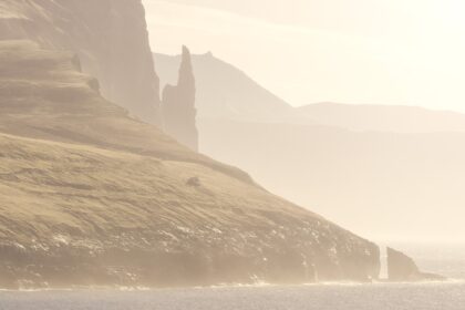 Trøllkonufingur sea stack at sunrise in the Faroe Islands, with warm light on rugged cliffs and ocean.