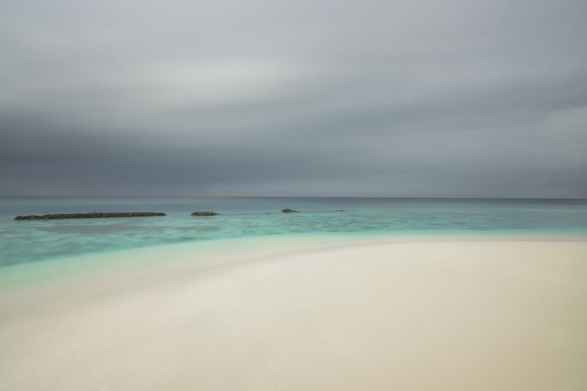 Long exposure landscape photography capturing a rainstorm on the paradise island of Thudufushi in the Maldives. Dark clouds swirl overhead as rain pours down, creating a dramatic and captivating scene.