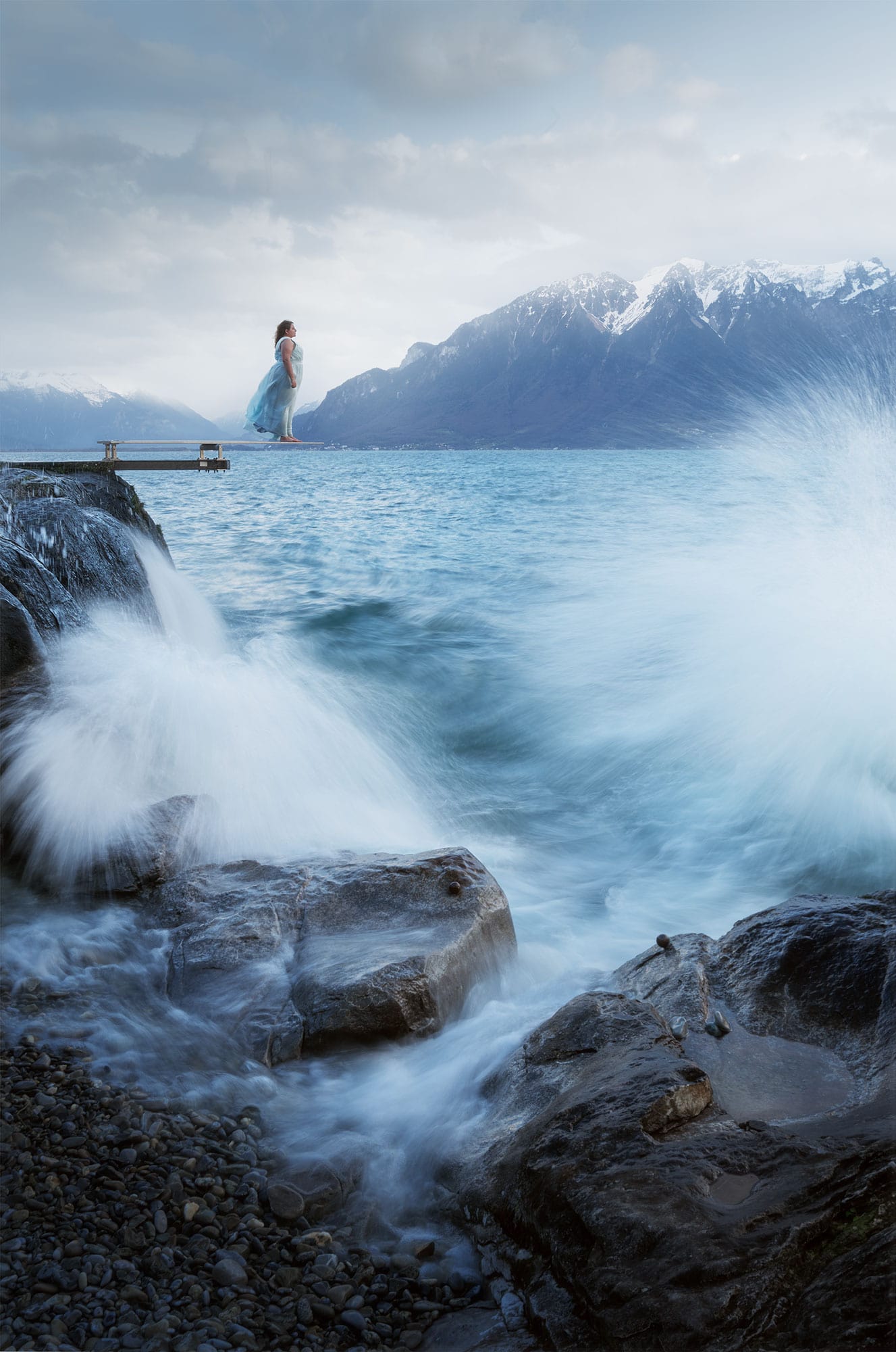 A self-portrait depicting a person wearing a turquoise princess gown, standing confidently on a diving board in the Lavaux region, overlooking Lake Geneva in Switzerland. The landscape photography captures the scene during the blue hour, with big waves crashing against the shore and the wind blowing fiercely. The figure gazes out at the vast expanse of the lake, surrounded by the stunning Swiss scenery.