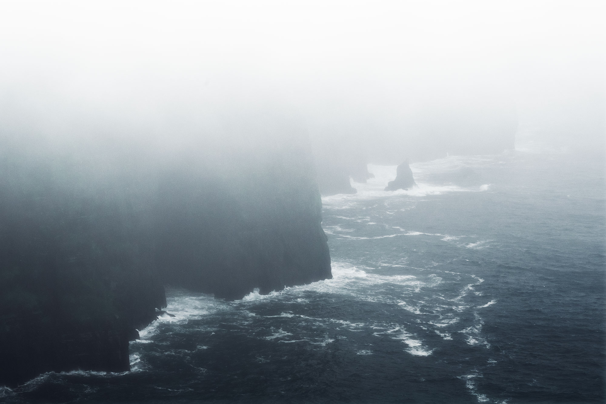 Epic and dramatic landscape photograph capturing the Cliffs of Moher in Ireland. The cliffs are enveloped in thick fog, creating a moody and mysterious atmosphere. The rugged coastline and towering cliffs are shrouded in mist, adding to the dramatic allure of the scene.
