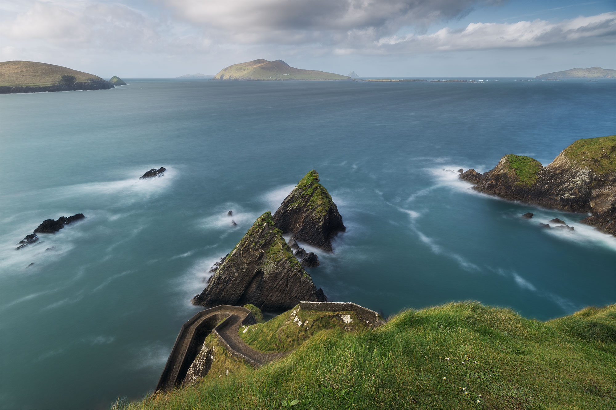 Jennifer Esseiva, a skilled photographer, captures the serene beauty of Dunquin Pier (Cé Dhún Chaoin) on the picturesque Dingle Peninsula along the Wild Atlantic Way in Ireland. Using her Nikon Z8, she employs a long exposure technique to depict the tranquil waters surrounding the pier, creating a mesmerizing effect. This landscape photograph invites viewers to immerse themselves in the tranquil ambiance of this iconic Irish coastal landmark.