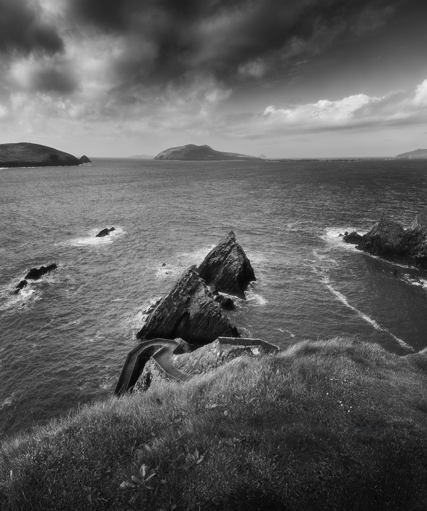 This striking black and white landscape photograph captures the timeless beauty of Dunquin Pier (Cé Dhún Chaoin) on the rugged Dingle Peninsula along the Wild Atlantic Way in Ireland. Taken by photographer Jennifer Esseiva using her Nikon Z8, the image employs a long exposure technique to evoke a sense of tranquility amidst the crashing waves. The contrast between light and shadow accentuates the dramatic contours of the pier, inviting viewers to contemplate the raw essence of this iconic Irish coastal landmark.