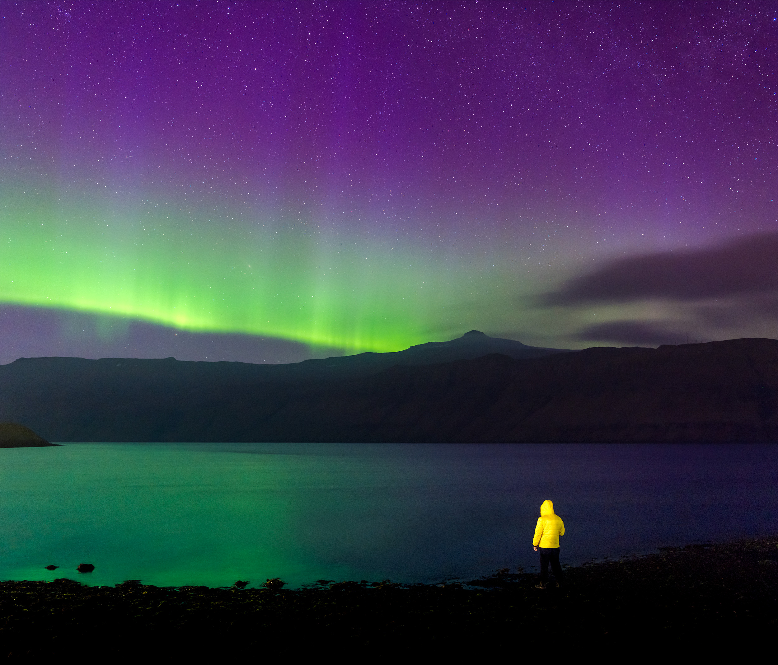 A landscape photograph capturing vibrant green and violet northern lights in the Faroe Islands. The image features a man wearing a yellow jacket, standing beneath the stunning aurora-filled sky.