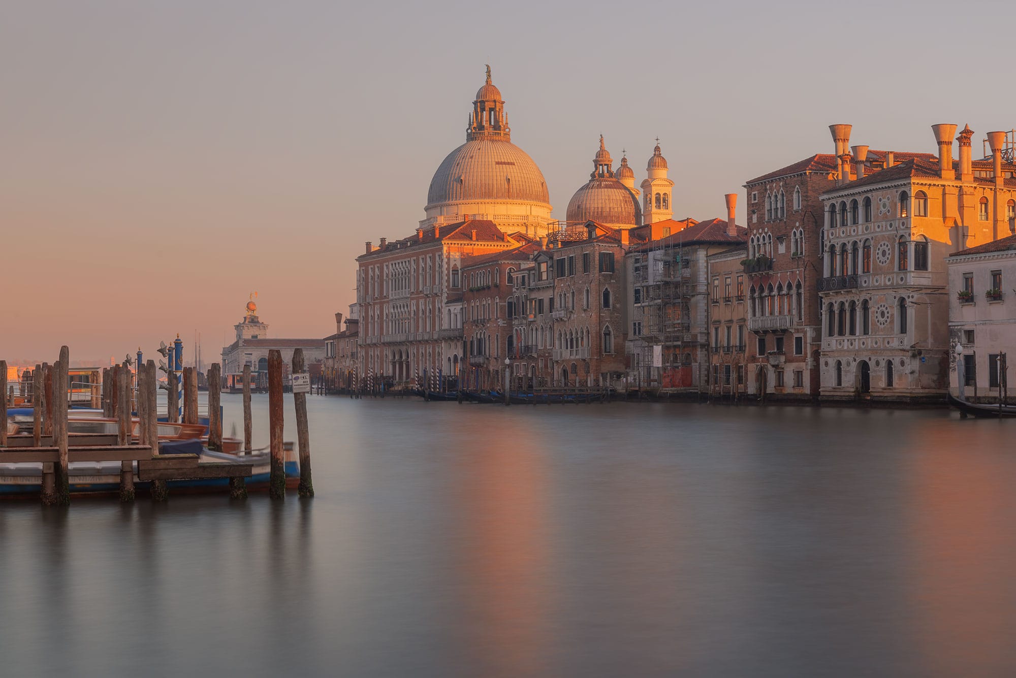 Urban photography capturing a long exposure of the Grand Canal in Venice, Italy, at sunset. The serene scene showcases the canal's smooth waters and the glowing hues of the setting sun.