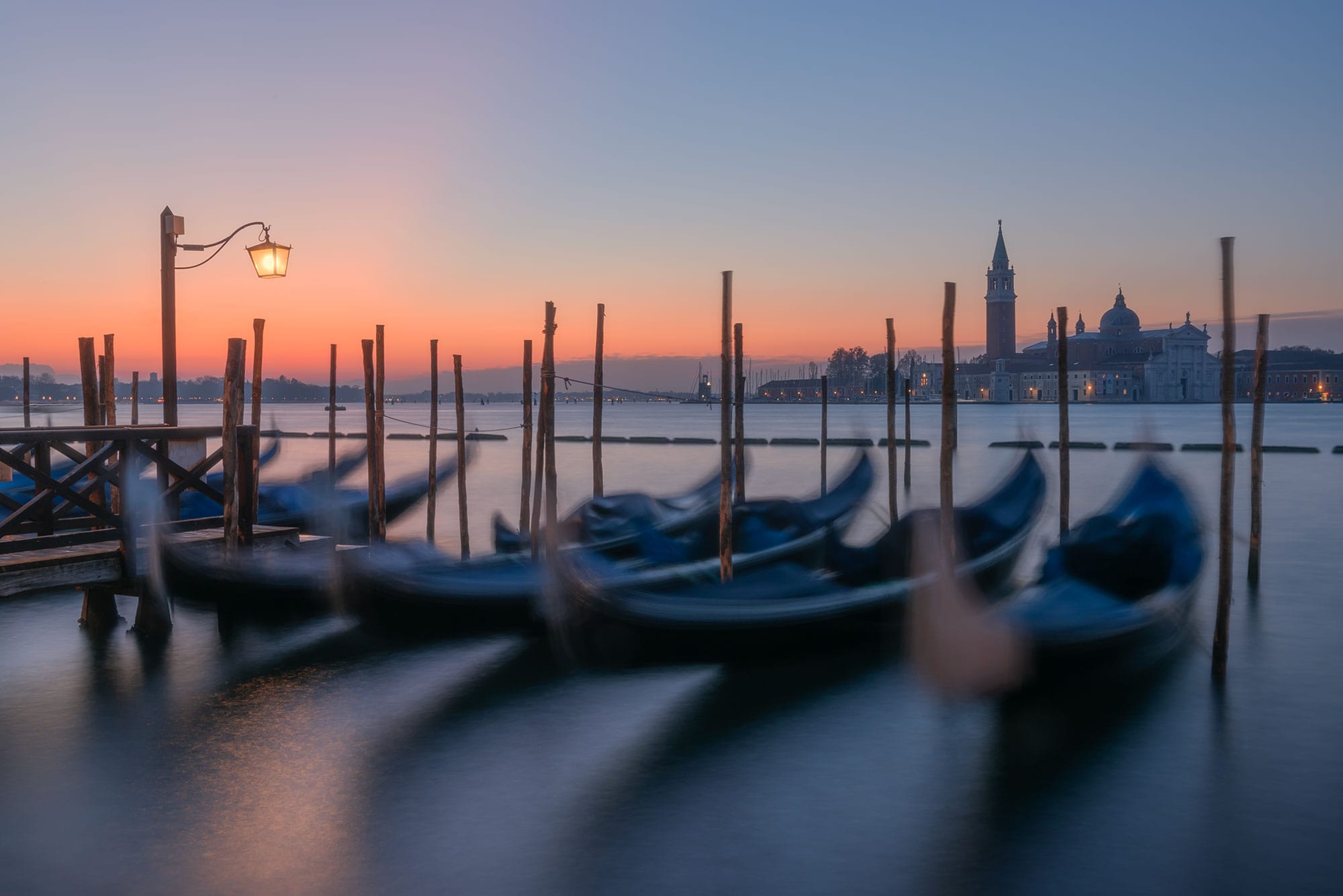Landscape photography capturing a sunrise at Piazza San Marco in Venice, featuring a long exposure shot of Venetian gondolas gently swaying on the water.