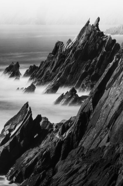 Black and white long exposure of black rocks at Coumeenoole Beach, Dingle Peninsula, Ireland, with rugged coastline and waves.