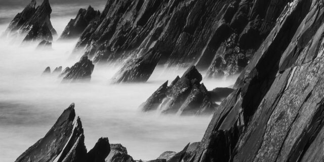 Black and white long exposure of black rocks at Coumeenoole Beach, Dingle Peninsula, Ireland, with rugged coastline and waves.