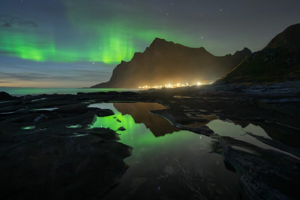 Landscape photography of the northern lights during the blue hour in the Lofoten Islands, Norway. Green aurora borealis glows above the rugged Arctic coastline, with rocky foreground textures and reflections under a deep blue sky.
