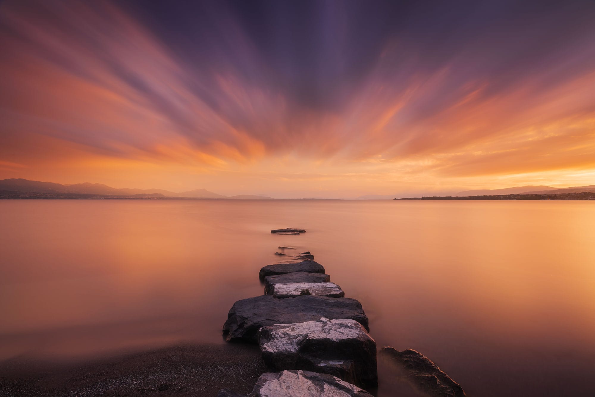 Landscape photography of Lake Geneva from Préverenges Beach at sunset, with long exposure and the Swiss Alps in the background.