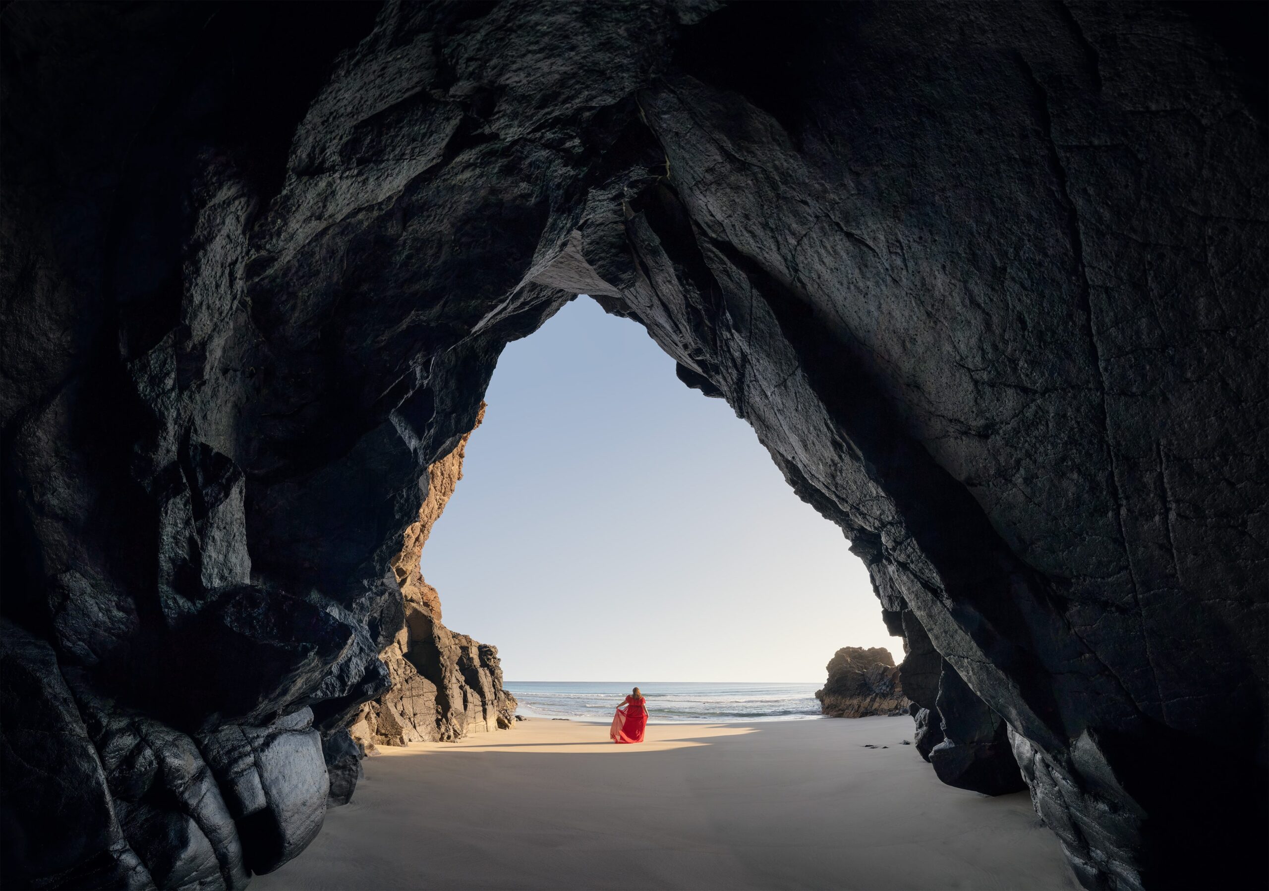 Panoramic self-portrait taken at sunrise on Órzola beach in Lanzarote, Canary Islands. The photographer, dressed in a flowing red dress, stands alone on the quiet black sand, framed by the volcanic cave's opening. The scene captures a moment of stillness and freedom, where soft morning light meets the raw beauty of the Atlantic coastline. Shot with a Nikon Z8, this image blends landscape, movement, and solitude into one timeless composition.