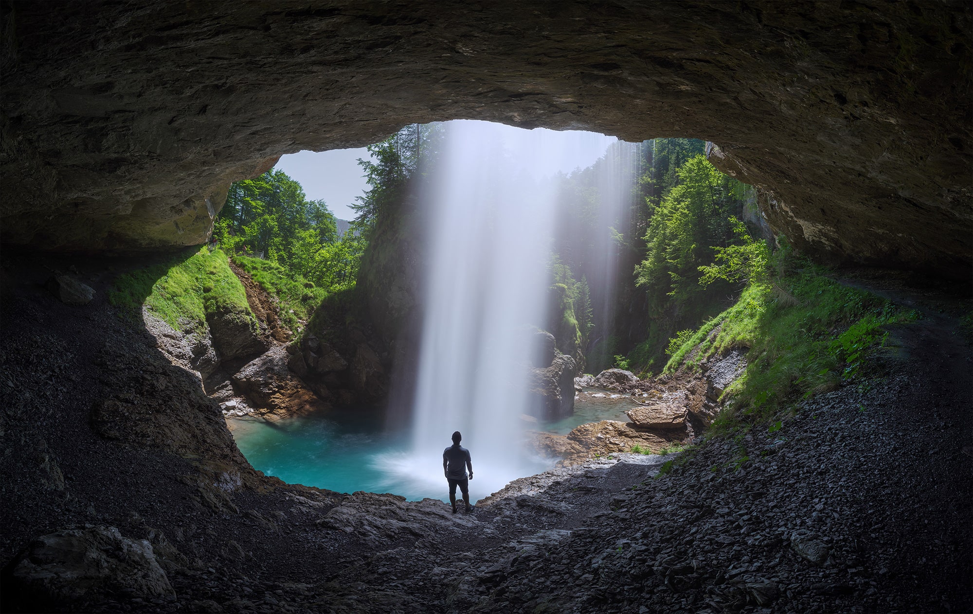 Panoramic long exposure photo of Bärglistüber Waterfall in the canton of Glarus, Switzerland, taken from behind the falls inside the cave. A person stands silhouetted in the scene, highlighting the scale of the natural rock formation. Captured with a Nikon Z8.