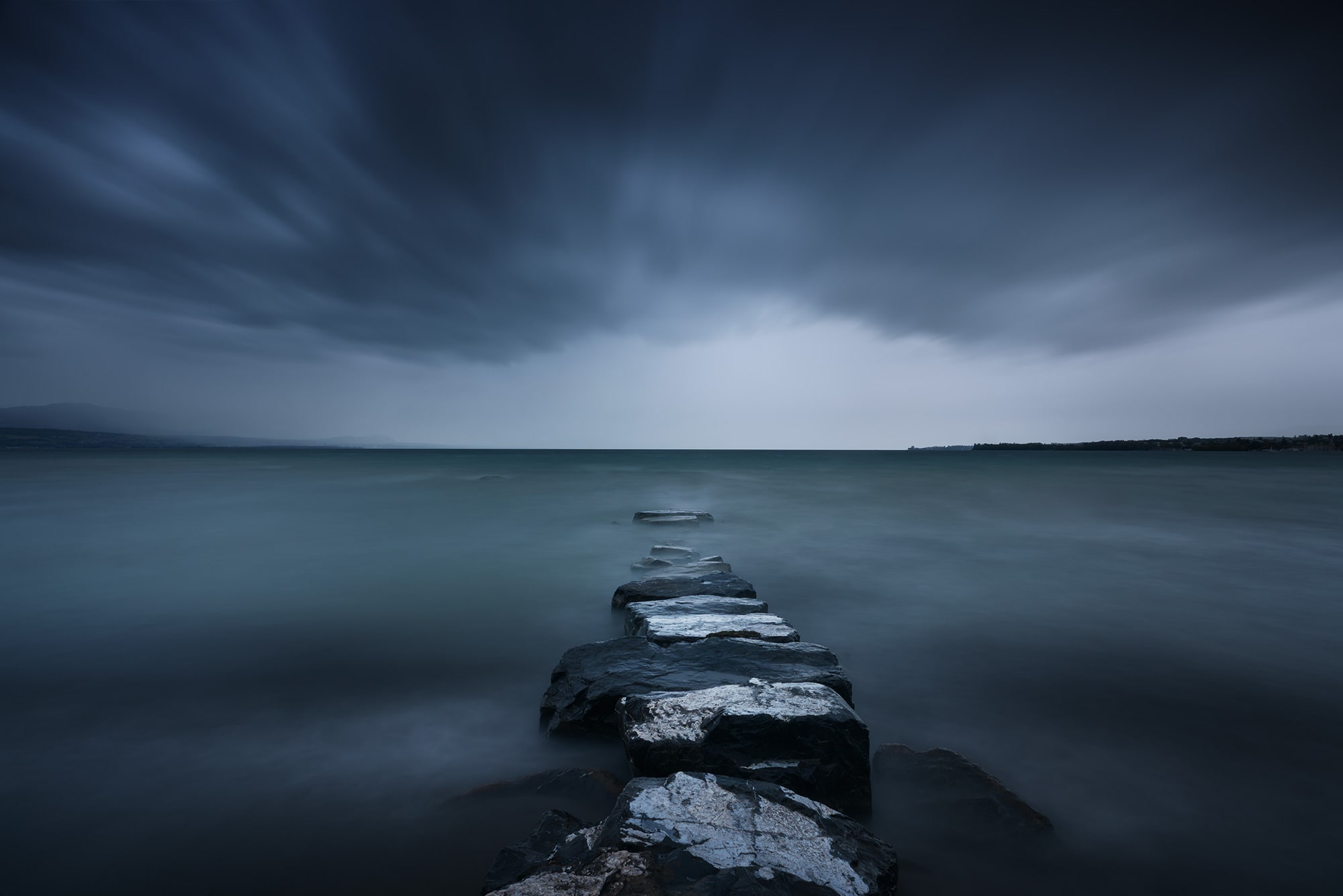 Epic sky landscape photography showing a dramatic storm approaching over Lake Geneva, captured from Préverenges beach near Lausanne, Switzerland. Pink sunset light contrasts with the incoming clouds.