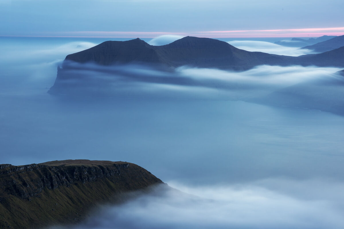 alt="Blue hour view from Sornfelli, Faroe Islands, with soft fog drifting over gentle mountain shapes. Captured with a long exposure, the image reveals muted tones and a quiet, intimate mood—offering a delicate alternative to the islands' usual dramatic atmosphere.