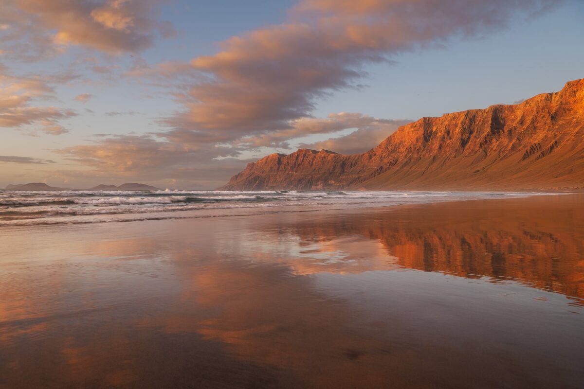 Sunset at Famara Beach in Lanzarote, Canary Islands, featuring soft light and a perfect reflection on the wet sand with mountains in the background.