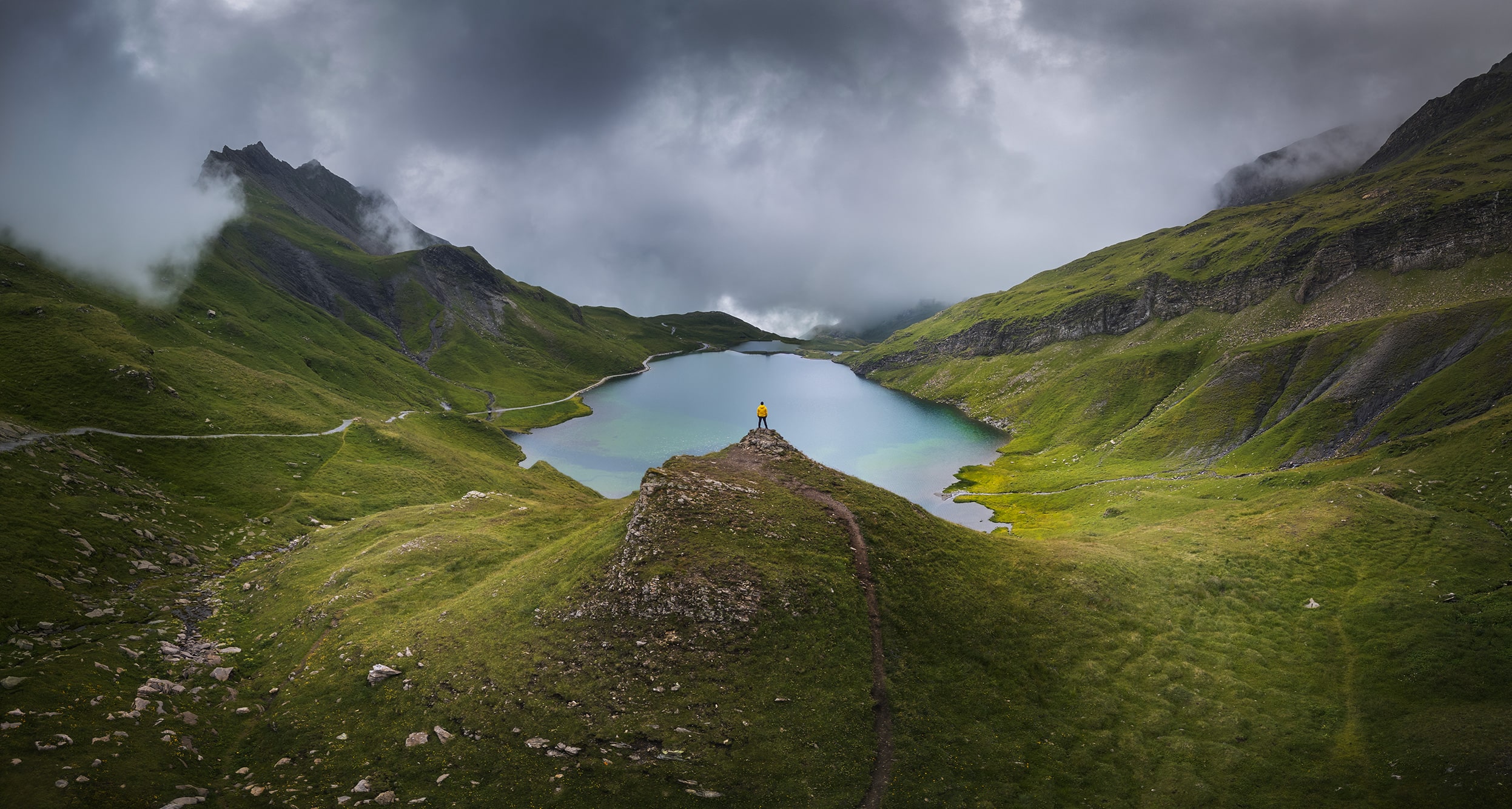 "Aerial landscape photography of Bachalpsee near Grindelwald, Switzerland, showing a wide panoramic view with a man in a yellow jacket admiring the alpine scenery.